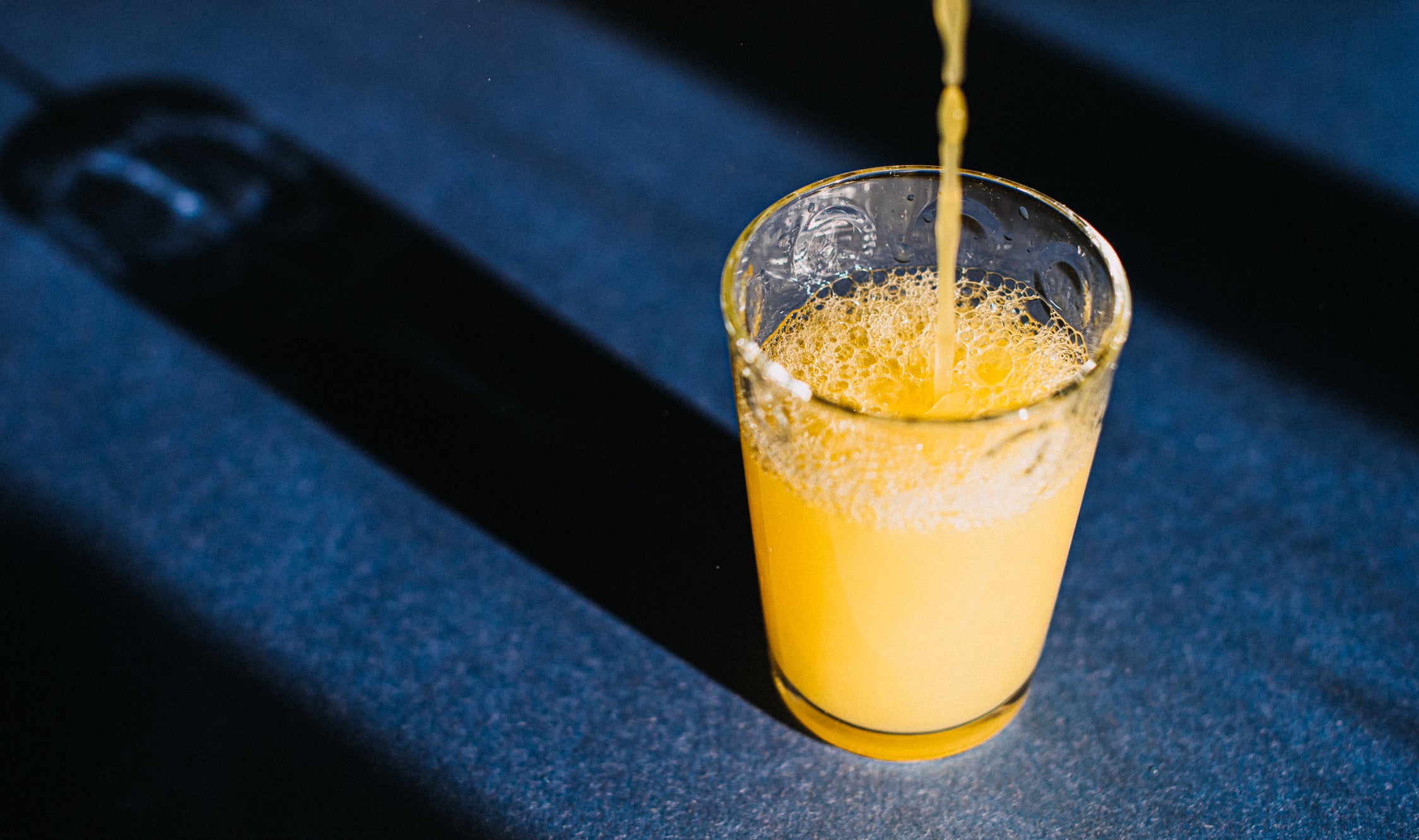 Orange Juice Being Poured in a Glass that casts a beautiful shadow on a blue surface.