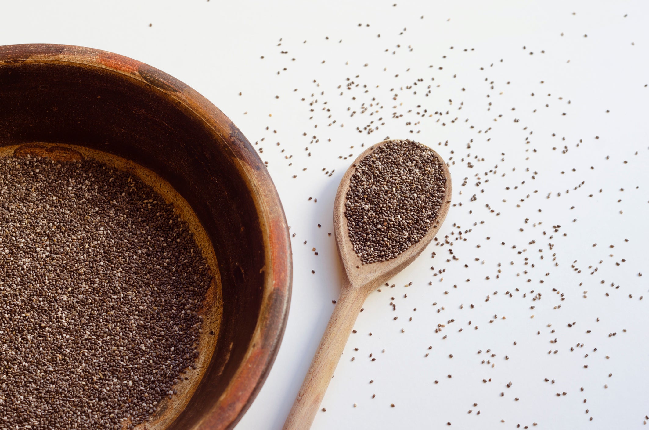 Clay vessel and wooden spoon with chia seeds on white background, seeds scattered on the table