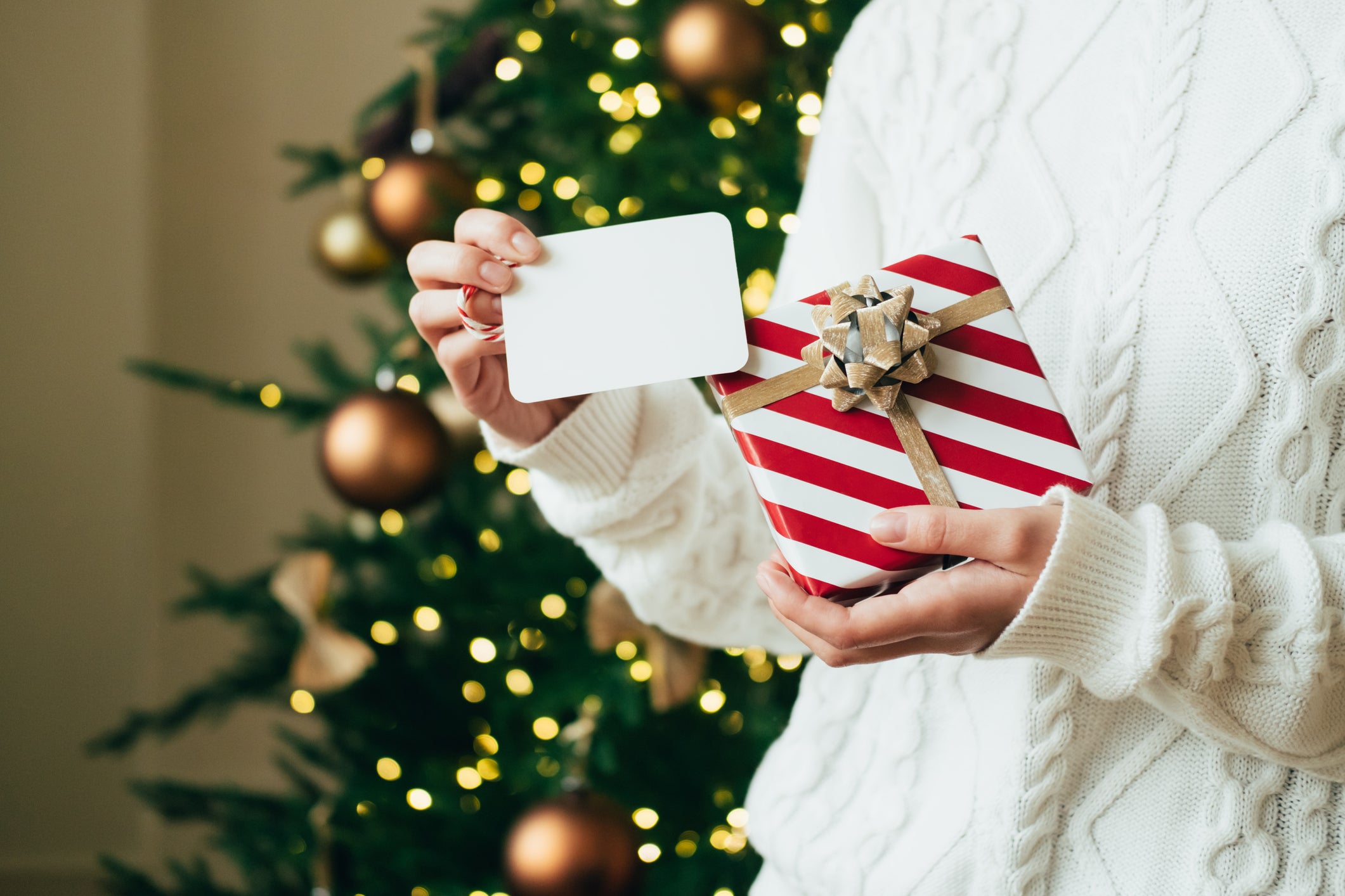 Woman in white sweater is holding red-white gift box with gold bow and white tag against lights of decorated Christmas tree. New Year celebration concept. Front view. Close-up